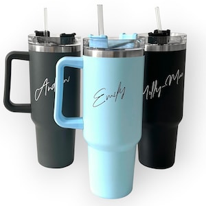 May include: Three personalized insulated tumblers with handles and straws. One is dark gray with "Andrew" in white script, another is light blue with "Emily", and the third is black with "Molly-Mae". All have silver lids.