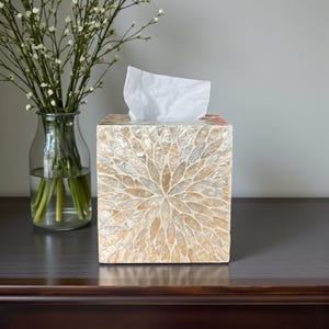 Könnte beinhalten: Ein quadratischer Taschentuchspender mit einem dekorativen, mosaikartigen Muster in Beige- und Cremetönen. Die Box hat ein Blumendesign und ein weißes Taschentuch wird von oben herausgezogen. Links steht eine Glasvase mit weißen Blumen.