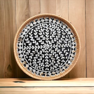 Puede incluir: Una bandeja redonda hecha de ratán tejido con un diseño de incrustaciones de nácar blanco y negro. La incrustación presenta un patrón circular de hojas blancas sobre un fondo negro.