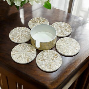 May include: Set of six round coasters with a white and gold iridescent design. The coasters are made of mother of pearl and have a floral pattern. The coasters come with a matching holder.