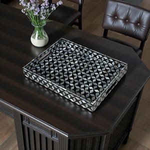 May include: A decorative rectangular tray with a black and white geometric pattern. The tray is made of a dark material and sits on a dark wooden table. A vase of purple flowers is in the background.