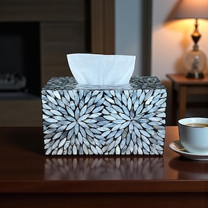 May include: A decorative tissue box with a mosaic design. The box is rectangular and covered in iridescent, gray-blue shell pieces arranged in a floral pattern. White tissues are visible at the top. The box sits on a dark brown surface.