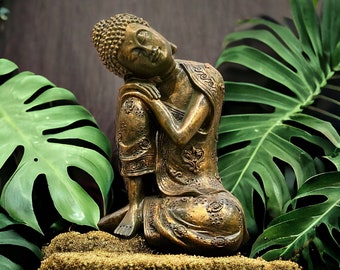 May include: A golden bronze statue of a meditating Buddha, sitting with his head resting on his hand. The statue is surrounded by lush green foliage.