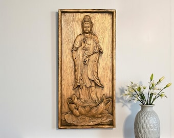 May include: A wooden relief carving of a standing Buddha figure, framed in a rectangular shape. The Buddha is depicted in flowing robes, standing on a lotus flower. The carving is in shades of brown and gold, mounted on a white wall.