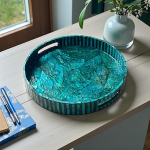 Könnte beinhalten: Ein runder dekorativer Tablett mit einer türkisfarbenen Mosaikoberfläche und schwarz gestreiften Kanten. Das Tablett hat zwei Griffe und steht auf einer hellen Oberfläche. Eine kleine weiße Vase mit Blumen befindet sich im Hintergrund.