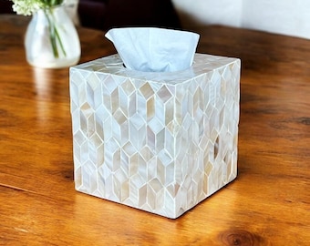 May include: A square tissue box with a mosaic pattern of white and beige shell pieces. The box has a geometric design and a single tissue is pulled from the top. The box is on a wooden surface.