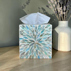 Puede incluir: Una caja para pañuelos cuadrada con un patrón de mosaico de piezas de concha iridiscentes en forma de pétalo en tonos azul claro, blanco y plata. Los fragmentos de concha están dispuestos en un diseño floral radiante, con un pañuelo blanco arriba.