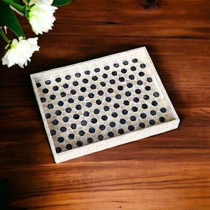 Può includere: Un vassoio decorativo rettangolare con un motivo a mosaico. Il bordo del vassoio è bianco e l'interno presenta un motivo ripetuto di piastrelle esagonali blu scuro e bianco sporco. Il vassoio è appoggiato su una superficie di legno scuro, con fiori bianchi sullo sfondo.