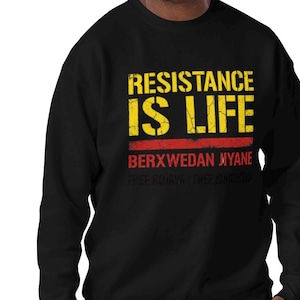 May include: Black crew neck sweatshirt featuring the words "RESISTANCE IS LIFE" in yellow, with a red stripe beneath. The text "BERXWEDAN JIYANE" and other white text are below. A casual, comfortable garment.