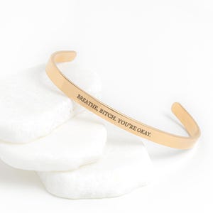 May include: A gold-colored cuff bracelet with the engraved text "BREATHE, BITCH. YOU'RE OKAY." The bracelet is resting on a stack of white, smooth stones. The bracelet has a simple, open design.