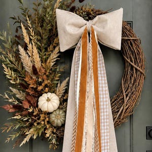May include: A circular grapevine wreath decorated with dried foliage, grasses, and two small pumpkins. A large beige fabric bow is attached to the top, featuring long, trailing ribbons in shades of burnt orange, cream, and a beige and white gingham pattern.