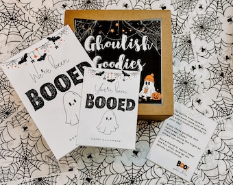 May include: A Halloween-themed gift set with a brown box and cards. The cards read "We've been BOOED" and "You've been BOOED" with ghost illustrations. The box says "Ghoulish Goodies". The background is a white fabric with black spiderwebs.