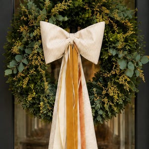 May include: A round wreath made of dark green boxwood leaves and sprigs of yellow foliage, featuring a large, centered bow made of beige and white striped fabric with long, hanging ribbons in mustard yellow, cream, and burnt orange velvet.