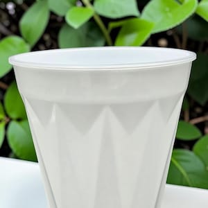May include: A white plastic flower pot with a geometric diamond pattern. The pot has a slightly flared rim and a smooth, clean surface. The background features green foliage.