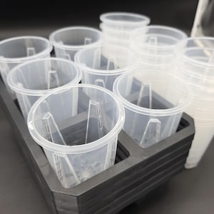 May include: A set of clear plastic plant pots with a black plastic tray. The pots are cylindrical with a flared rim and internal supports. The tray has multiple compartments to hold the pots.