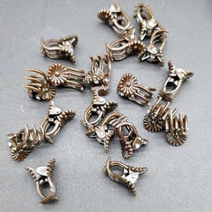 May include: A collection of bronze-colored metal hair clips. The clips feature a variety of designs, including floral and geometric shapes. The clips are scattered on a gray surface, showcasing their intricate details and metallic sheen.