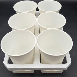May include: A white plastic cup holder with six white paper cups. The cups are empty and have a rounded shape. The holder has a rectangular shape with handles.