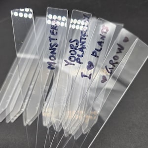 May include: A set of clear plastic plant markers with black handwritten labels. The labels read "Monster", "Yours Plants", "I ♥ Plants", and "Grow ♥".