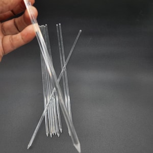 May include: A collection of clear, pointed plastic rods. One is held in a hand, while others are scattered on a dark surface. The rods appear to be thin and transparent, with a sharp, conical tip.