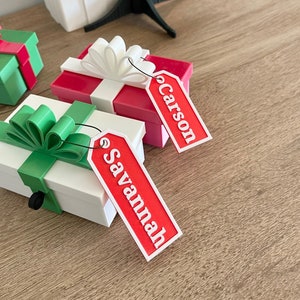 May include: Two small gift boxes with red and white ribbon and green bows. The boxes have personalized tags with the names "Carson" and "Savannah" written in white.