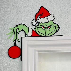 May include: A green Grinch-themed door frame decoration. The Grinch is wearing a red Santa hat and red and white outfit, with a red ornament hanging from his hand. The decoration is placed on a white door frame.