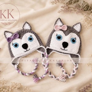 May include: Two handmade crochet hats designed to look like husky dogs. Each hat is grey and white with blue eyes, a black nose, and ear flaps with braided tassels. One hat features a purple bow, and the other features a light pink bow.