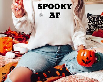 May include: A white sweatshirt with the text "SPOOKY AF" in black letters. A small spider is hanging from the "Y" in "SPOOKY". The model is wearing the sweatshirt and blue jeans.