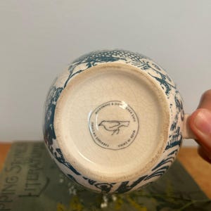Op de afbeelding: Een witte keramische mok met een blauwgroen bloemmotief. De bodem van de mok heeft een ronde stempel met een vogelillustratie en de tekst "Microwave & Dishwasher Safe Made in China". De mok wordt door een hand vastgehouden.