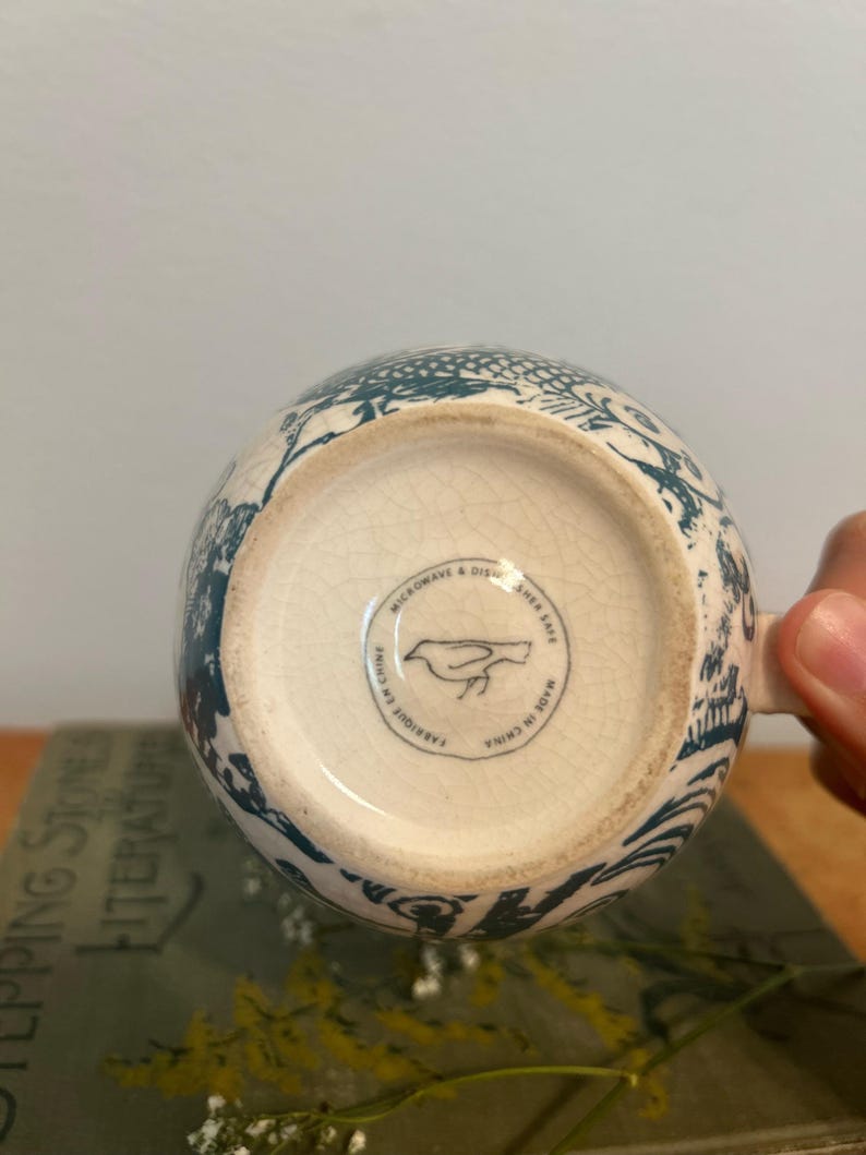 Op de afbeelding: Een witte keramische mok met een blauwgroen bloemmotief. De bodem van de mok heeft een ronde stempel met een vogelillustratie en de tekst "Microwave & Dishwasher Safe Made in China". De mok wordt door een hand vastgehouden.