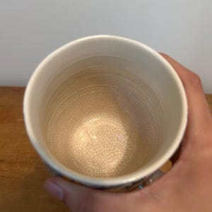 Op de afbeelding: Een witte keramische mok met een getextureerd interieur en een licht uitlopende rand. De mok heeft een craquel&eacute;-glazuureffect aan de binnenkant en wordt door een hand vastgehouden. De mok staat op een houten oppervlak.
