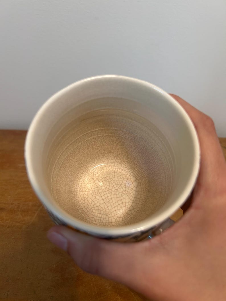 Op de afbeelding: Een witte keramische mok met een getextureerd interieur en een licht uitlopende rand. De mok heeft een craquel&eacute;-glazuureffect aan de binnenkant en wordt door een hand vastgehouden. De mok staat op een houten oppervlak.