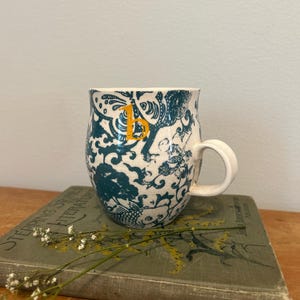 Op de afbeelding: Een witte keramische mok met een blauwgroen en wit patroon en een gele letter "B". De mok heeft een gebogen vorm en een wit handvat. De mok staat op een vintage boek met de titel "Stepping Stones to Literature".