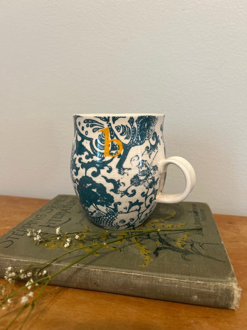 Op de afbeelding: Een witte keramische mok met een blauwgroen en wit patroon en een gele letter "B". De mok heeft een gebogen vorm en een wit handvat. De mok staat op een vintage boek met de titel "Stepping Stones to Literature".