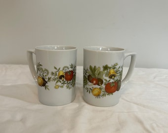 May include: Two white ceramic mugs with a floral design featuring vegetables, mushrooms, and bells. The mugs have a handle on the right side.