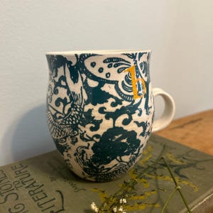 Op de afbeelding: Witte keramische mok met een teal en wit draken- en bloemenpatroon. De mok heeft een wit handvat en een kleine gele letter "B" aan de zijkant. De mok staat op een vintage boek.