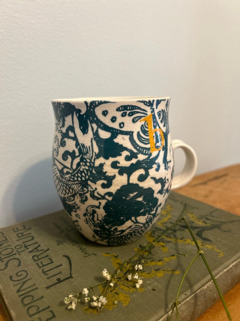 Op de afbeelding: Witte keramische mok met een teal en wit draken- en bloemenpatroon. De mok heeft een wit handvat en een kleine gele letter "B" aan de zijkant. De mok staat op een vintage boek.
