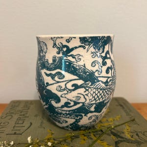 Op de afbeelding: Een keramische beker met een ontwerp van turquoise koi-karpers. De vissen zwemmen tussen wervelende patronen tegen een witte achtergrond. De beker staat op een boek met de titel "Living Stones to Literature".
