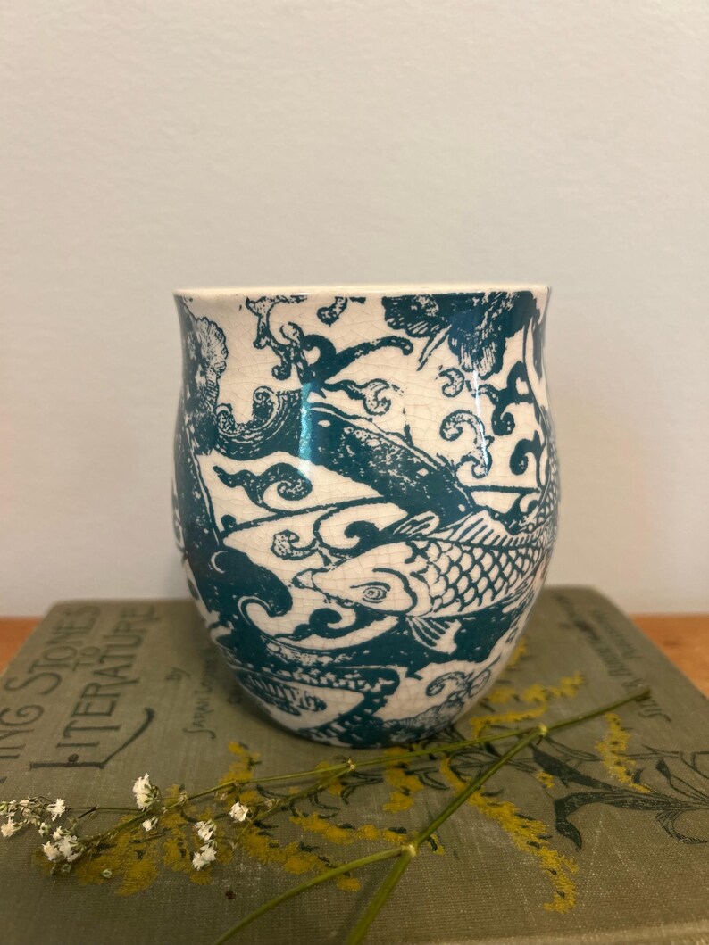 Op de afbeelding: Een keramische beker met een ontwerp van turquoise koi-karpers. De vissen zwemmen tussen wervelende patronen tegen een witte achtergrond. De beker staat op een boek met de titel "Living Stones to Literature".