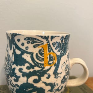 Op de afbeelding: Cr&egrave;mekleurige keramische mok met een teal en wit bloemenpatroon. De mok heeft een oranje letter "b" en een gebogen handvat. Het ontwerp omvat gestileerde bloemen en andere organische vormen. De mok staat op een groen boek.