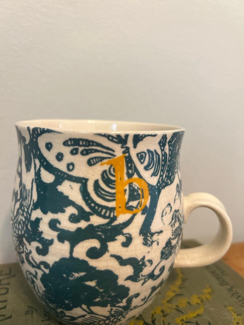 Op de afbeelding: Cr&egrave;mekleurige keramische mok met een teal en wit bloemenpatroon. De mok heeft een oranje letter "b" en een gebogen handvat. Het ontwerp omvat gestileerde bloemen en andere organische vormen. De mok staat op een groen boek.