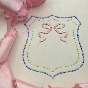 May include: A white embroidered shield design with a pink bow in the center. The shield is outlined in blue and green stitching. Pink ribbon with pom-pom trim is draped around the design.