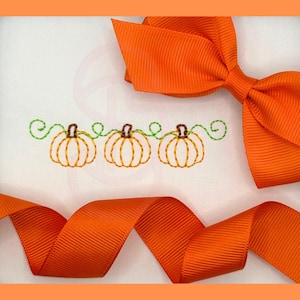 May include: A white fabric surface featuring an embroidered design of three orange pumpkins with brown stems and green swirling vines, accented by a bright orange grosgrain ribbon tied in a bow at the top and draped across the bottom.