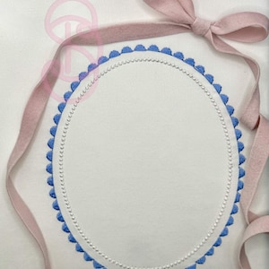 May include: White fabric bib with an embroidered oval design. The oval is outlined with blue scalloped edges and a line of white beads. A pink ribbon is tied at the top, and the bib has pink ribbon ties.