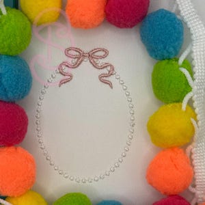 Puede incluir: Un diseño ovalado enmarcado en blanco con un lazo rosa y un borde de cuentas. El marco está rodeado de pompones coloridos en tonos de amarillo, verde, naranja, rosa y azul. Una cinta blanca con un borde decorativo está en el lado derecho.