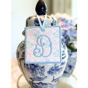 May include: A square, embroidered tag with a light blue border and a light blue letter "D" in a script font. The tag has a pink and white floral pattern and is suspended by a white ribbon. The tag is hanging on a blue and white porcelain jar.