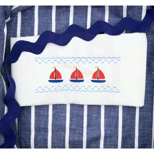 May include: White rectangular pillowcase with an embroidered design of three red sailboats with blue hulls and yellow flags. The pillowcase is set on a blue and white striped fabric, with a wavy blue ribbon.