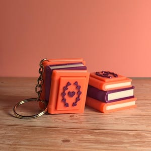 May include: Orange and purple miniature book keychains. One keychain has a silver ring and chain. The books have white pages and a decorative design on the cover. One stack of books is on the right.