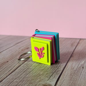 May include: A stack of colorful mini-books attached to a silver keyring. The books are neon green, pink, white, and teal. The front book has a pink floral design. The background is a light pink.