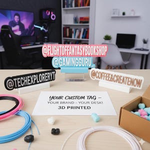 Op de afbeelding: Een verzameling 3D-geprinte aangepaste bureaubordjes met social media handles, waaronder @FLIGHTOFFANTASYBOOKSHOP, @GAMINGGURU, @TECHEXPLORERYT en @COFFEE&CREATENOW. De bordjes liggen op een bureau met 3D-printfilament en een kartonnen doos.