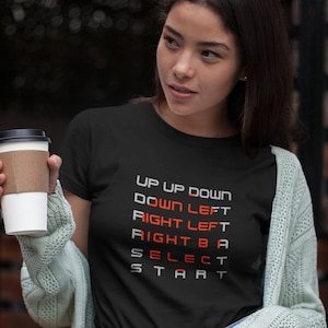 May include: A woman wearing a black t-shirt with the words "UP UP DOWN DOWN LEFT RIGHT LEFT RIGHT B A SELECT START" in red and white text. She is holding a brown paper cup in her left hand and wearing a light green cardigan and blue denim shorts.